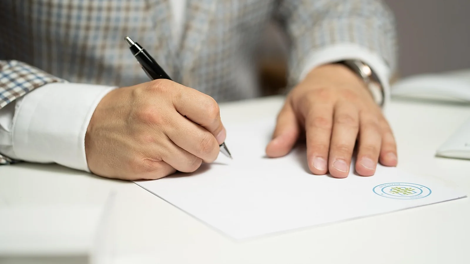 A person signs a printed document with a pen at a white desk