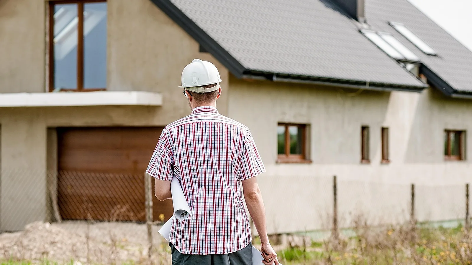 A person in a hard hat carrying rolled plans walks toward a house