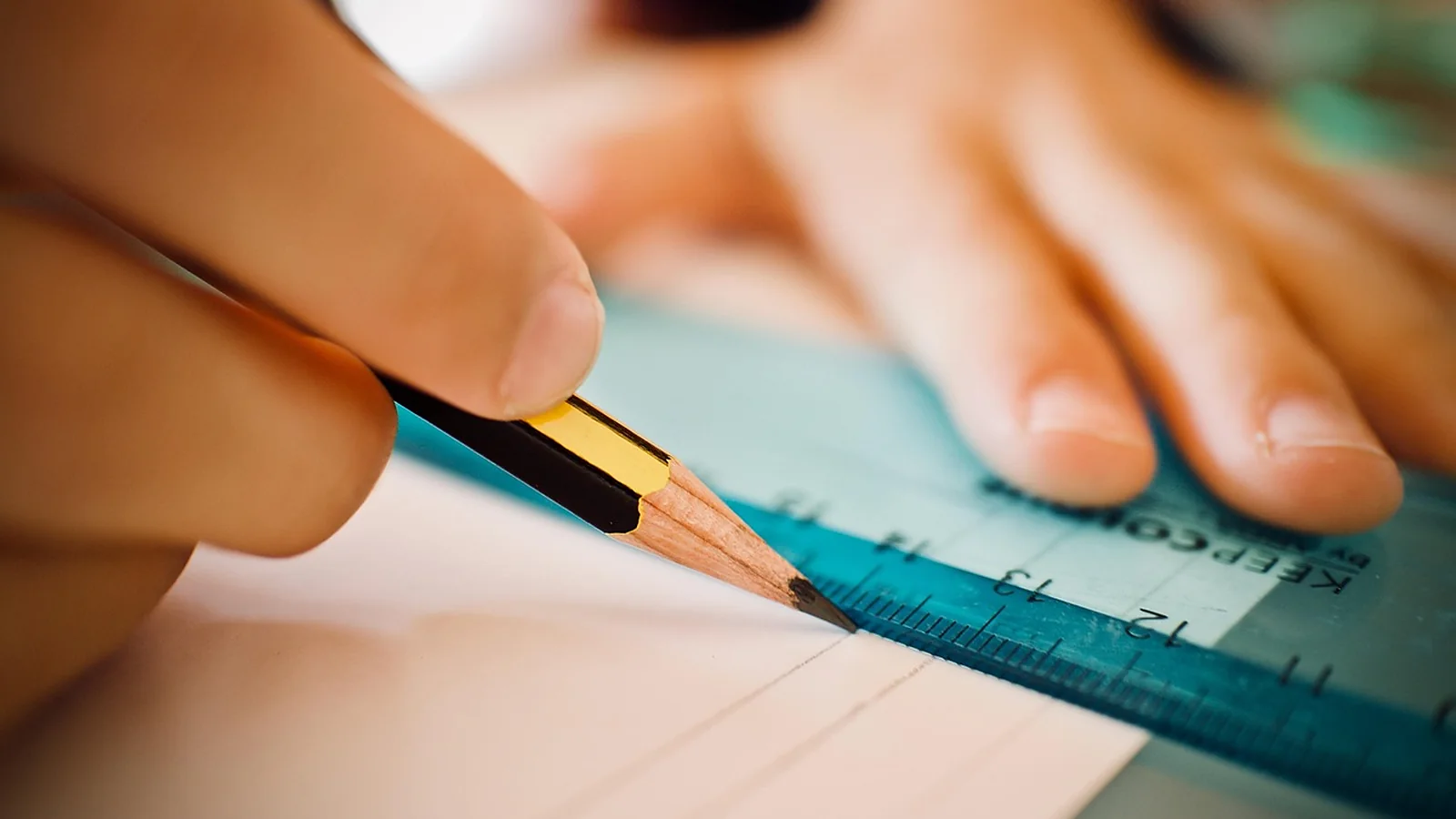A hand draws a straight line with a pencil and ruler on a sheet of paper