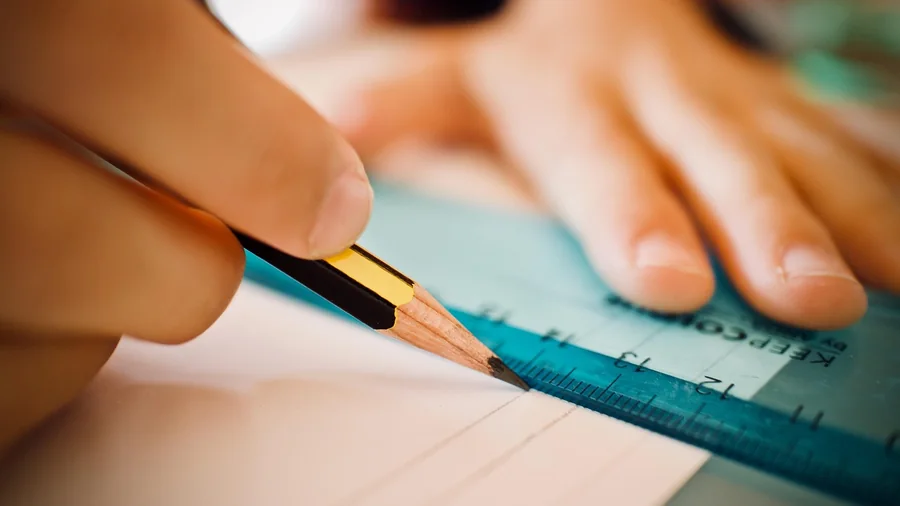 A hand draws a straight line with a pencil and ruler on a sheet of paper
